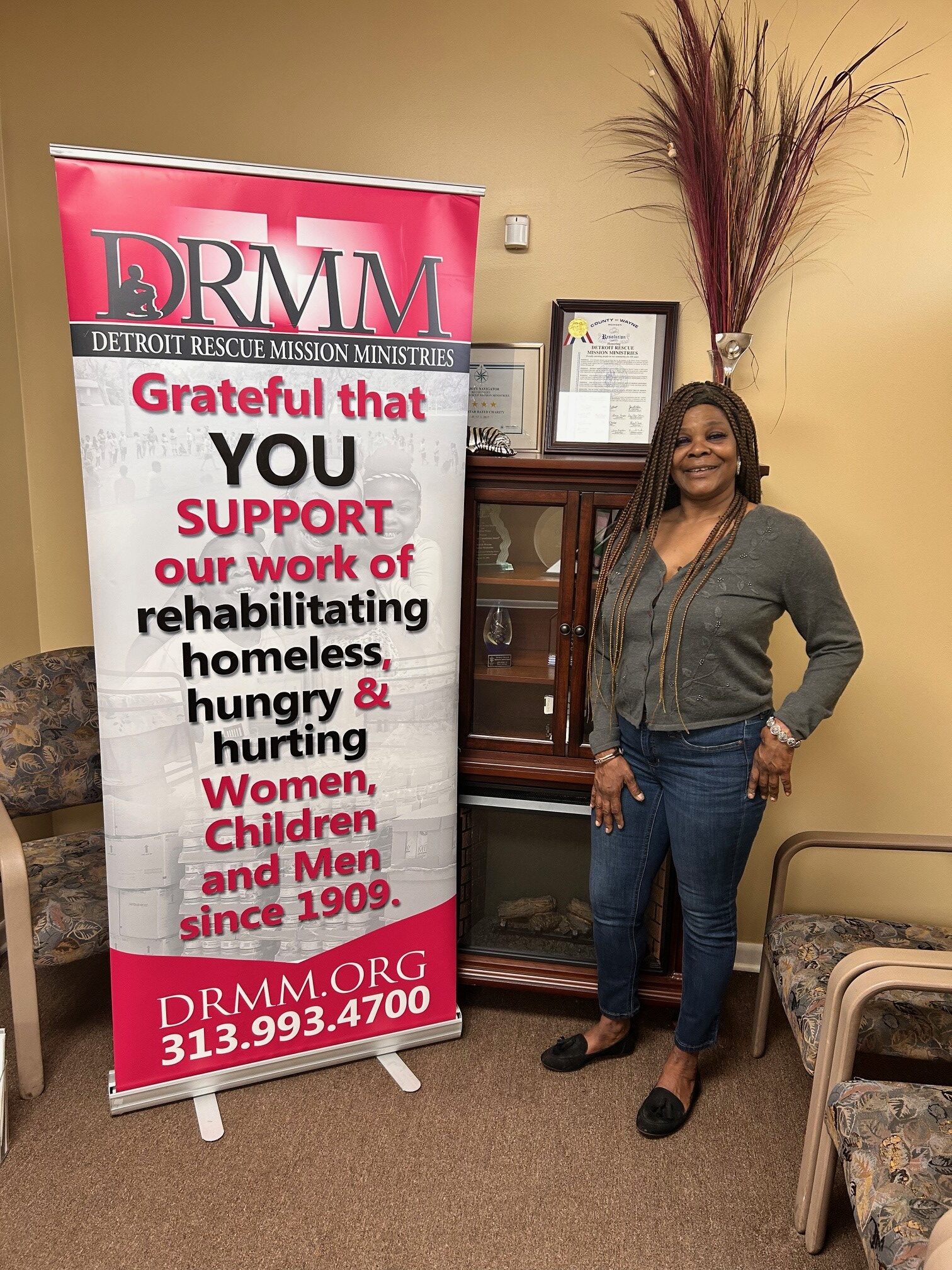 This is Martina standing in a gathering room next to a sign that reads, "DRMM grateful that YOU support our work of rehabilitating homeless, hungry & hurting women, children and men since 1909."